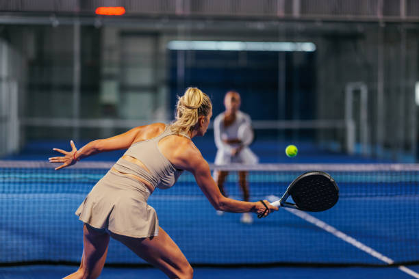 Rear view of a blonde Caucasian female padel player hitting a forehand with a black padel racket in a singles padel match.